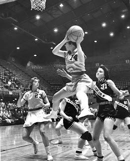 Black and white action photograph of a women's college basketball game, a player driving to the basket in a packed arena.