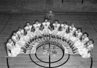 Mid-century women's basketball team photographed in a gymnasium, seated in a fan formation around a trophy at center court.